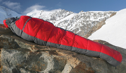 A photograph shows a person wrapped in a sleeping bag lying on rock high in snow-covered mountains. 
