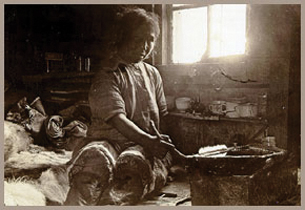 A black and white photograph shows an Inuit woman in a very basic house. The woman is sitting in front of a seal oil lamp—a semi-circular bowl containing seal oil. A row of small flames appears at the straight edge of the bowl.