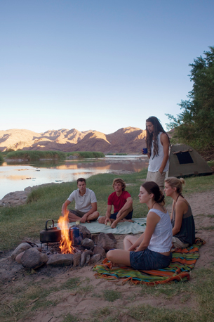 A photograph shows three men and two women sitting around a campfire. 