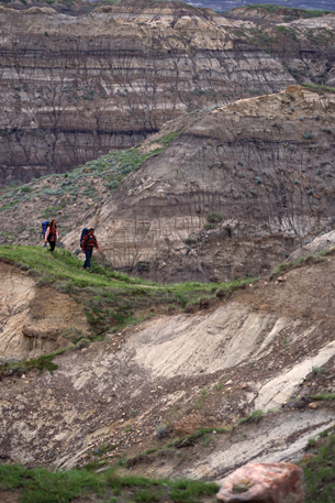 A photograph shows two hikers walking along a trail. The trail they are walking on briefly dips downward but then slopes upward again.