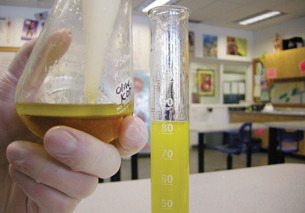 A photograph shows a flask with two layers: a brown layer on the bottom and a cloudy, yellow layer (biodiesel) on top. A hand is holding the flask and transferring the cloudy, yellow layer to a graduated cylinder sitting on a desk. 
