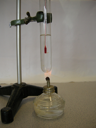 Photo 12: This photograph shows the test tube in the test-tube clamp. A thermometer is in the test tube and a spirit burner filled with methanol is beneath it. 
