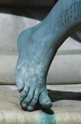 A photograph shows a copper statue of a foot. The statue has turned green; the green appears to have dripped onto the statue's stone base.