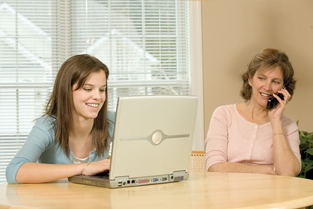 A photograph shows a high school-aged girl working on a laptop computer at a kitchen table. Her mother is sitting beside her and talking on a cordless telephone.