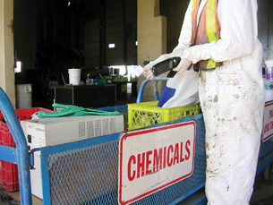 A photograph shows a person wearing white coveralls and protective gloves sorting discarded electronic materials at a disposal facility for special waste. The person in coveralls has a cordless telephone in one hand and the telephone’s rechargeable battery in the other. These items are being placed into separate areas of a large blue bin labeled “Chemicals.” A discarded computer and other electronic equipment are in the bin.