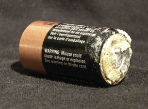 A photograph shows a commercial voltaic cell on its side. Solid crystalline material coats the outside of the cell at its flat end. Along the length of the cell is the following text: "Warning: Misuse could cause leakage or explosion. See warning on blister card."