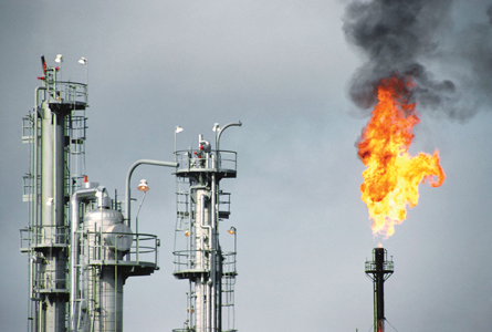 A photograph shows part of an oil refinery. The tops of three fractionating towers and a flare are shown. A bright orange flame is shown at the top of the flare. A cloud of thick black smoke rises above the flame.