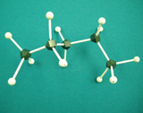 A photograph shows a molecular model for pentane, a form of C5H12. Black balls represent carbon atoms and white balls represent hydrogen atoms. All bonds between balls are a single connection. The model has five black balls joined together to form a chain. Three white balls are joined to the first and fifth black balls and two white balls are attached to each of the second, third and fourth balls. 
