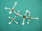 A photograph shows a molecular model for 2 –methylpentane, a form of C5H12. Black balls represent carbon atoms and white balls represent hydrogen atoms. All bonds between balls are a single connection. The model has four black balls joined together to form a chain. Three white balls are joined to the first and fourth black balls, and one white balls is attached to the second black ball. The remaining black ball with three white balls attached to it is attached to the second black ball in the chain. Two white balls are attached to the third black ball in the chain.