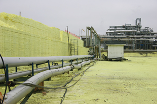 A photograph shows a facility that removes sulfur from natural gas. The facility is a multi–storey building with many exposed pipes. In the foreground of the picture is an extremely large yellow rectangular block of sulfur.