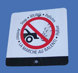 A photograph shows a road sign nailed to a blue background. The centre of the sign is a drawing of the back half of a car with a cloud of exhaust coming out its tailpipe. A red circle with a line across it is placed over the image of the car and the exhaust. Words in French and English appear above and below the image. The English words read “Stop•IDLING• Pollutes.”