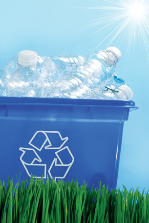 A photograph shows a blue, plastic box filled with empty, clear plastic water bottles sitting on grass. On the side of the box is the recycle logo—three arrows curved to form a triangle.
