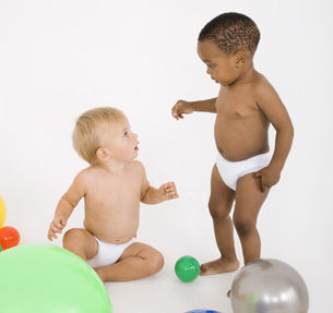 This is a photograph of two toddlers dressed in disposable diapers. One is sitting on the floor, while the other is standing. They have colourful toys to play with in the room, such as balls.