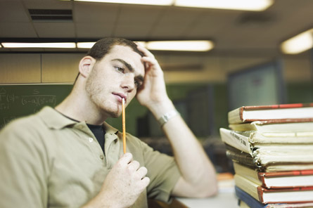 A photograph shows a man sitting in front of a pile of books. He is holding a pencil to his chin as though in deep thought. 