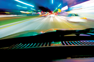 A photograph shows a blur of lights out the front window of a vehicle that is passing brightly lit objects and other vehicles while driving at night.