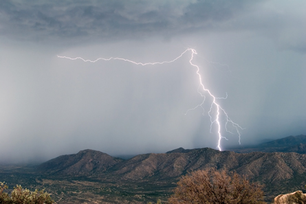 A photograph shows a lightning strike passing between a cloud and a mountain ridge.
