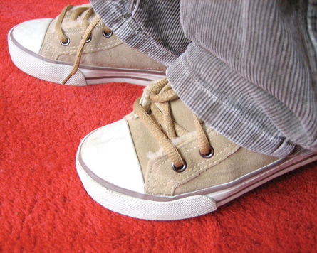 The photo shows a close-up of someone’s feet on a floor, with the hems of the person’s jeans drooping over the shoes. The shoes are tan coloured running shoes, with laces, white rubber toes and soles, with blue pinstripes along the soles.