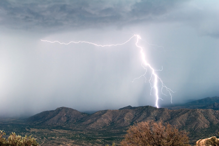 The photo shows a stormy sky above a mountainous ridge. Lightning is shown streaking across the sky and stretching from sky to ridge top. There are trees in the foreground.