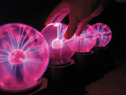 Four plasma balls are arranged in a line with a hand touching the second ball. The streamers in the second ball all go to the fingers on the hand. The other three have random streamers throughout.