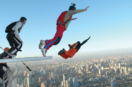 In this photo, three BASE jumpers wearing parachutes are shown leaping from the top of a tall structure. One of the jumpers appears to have a camera on his head. A city landscape is shown in the background.