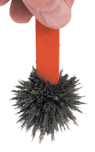 A photograph shows tiny iron filings clinging to the end of a red bar magnet held in someone’s hand. The filings reveal the shape of the magnetic field near the pole of the bar magnet.