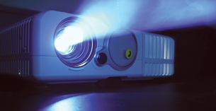A photo shows a film projector with a beam of bright light shining from it. Projection systems use digital light processing to produce fluid, vibrant images on a nearby screen.