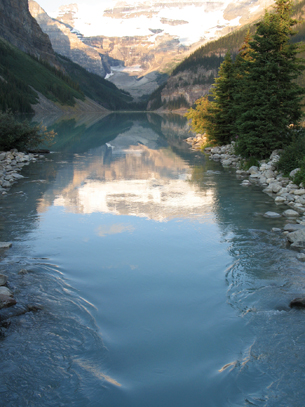 A photo shows a mountain in the background. In the foreground is a wide stream in which there is an inverted reflection of the mountain in the centre where the water is still.