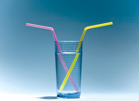 A photograph shows a clear glass of water with two tall straws standing in it. Refraction causes these partially submerged objects to appear broken or disconnected.