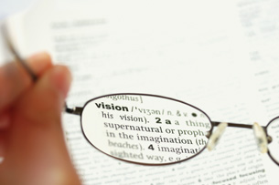 A photograph shows a pair of glasses held in front of a page in a dictionary. The text on the page in the background is blurry, but the text seen through one of the lenses is clear, and shows the definition of “vision.” The image shows that the lenses in a pair of glasses correct some vision problems by refracting light toward specific regions of the human retina.