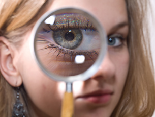 A photograph shows a woman holding a magnifying glass up in front of her right eye. Her eye appears very large within the magnifying glass. It shows that a large, virtual image is formed when a converging lens, such as the magnifying glass, is placed near an object.