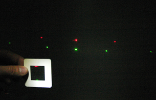 A red and green laser are shone on a diffraction grating and the maximum pattern can be seen on the screen. The red laser light pattern is wider than the green laser light pattern on the screen.