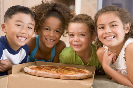 This photo shows four young children, two boys and two girls, gathered around a pizza.
