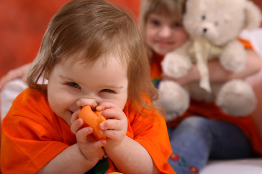 A developmentally challenged child plays with a small toy while an older child with a stuffed animal looks on.