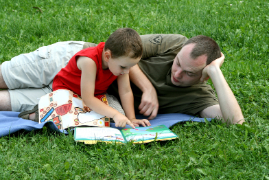 This photo shows a man and a small boy reading in an outdoor setting.