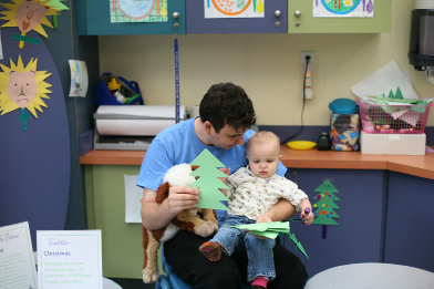 This photo shows a man holding a baby on his knees. They are looking at a variety of shapes cut out like trees.