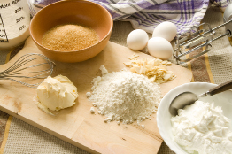 This photograph shows baking ingredients (eggs, flour, sugar, butter, and milk) on a table.