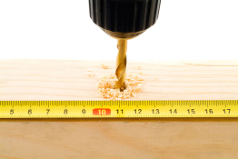 This is a photograph of a drill boring a hole in wood. A tape measure is extended next to the wood.