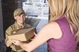 This photograph shows a female courier delivering a parcel to a woman at the door of her home.