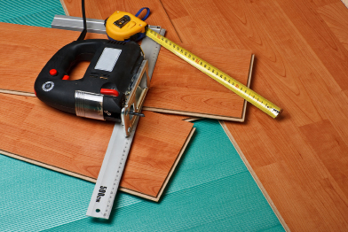 This photograph shows the installation of laminate flooring. A sabre saw, tape measure, and a square rest on two pieces of laminate flooring.