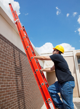 This is a photograph of a construction worker climbing a ladder that is propped against a flat roof.
